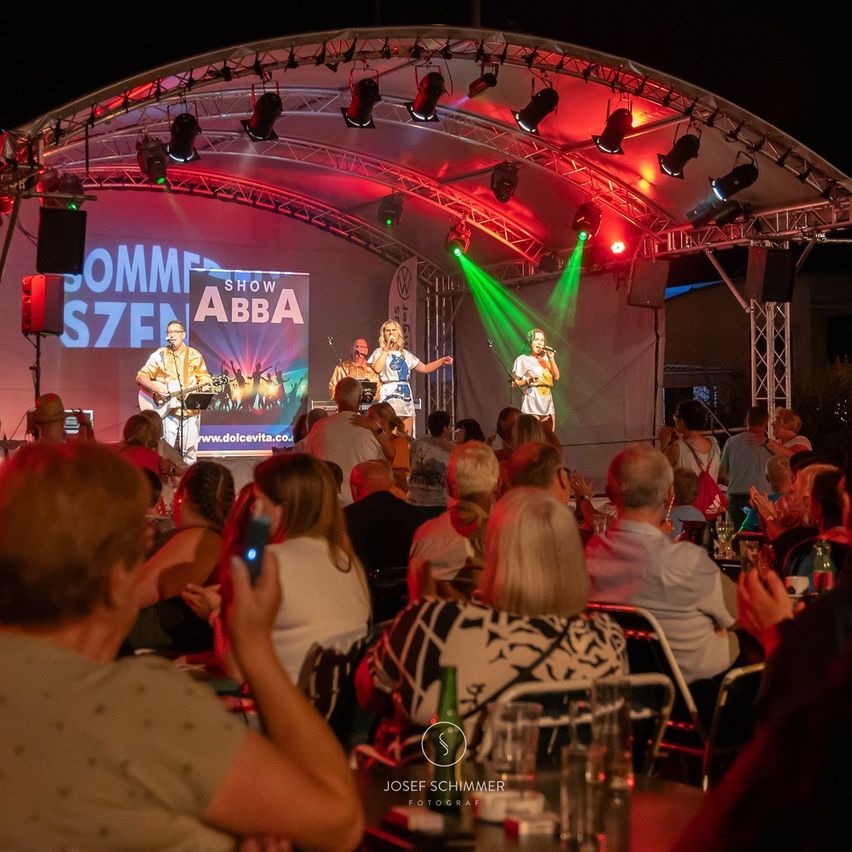 Eine Gruppe tritt bei Nacht auf der Bühne unter bunten Lichtern auf. Das Publikum sitzt davor, einige klatschen. Ein Banner mit 'SHOW ABBA' ist zu sehen. Das Bild hat einen Wasserzeichen von 'JOSEF SCHIMMER FOTOGRAF'.
