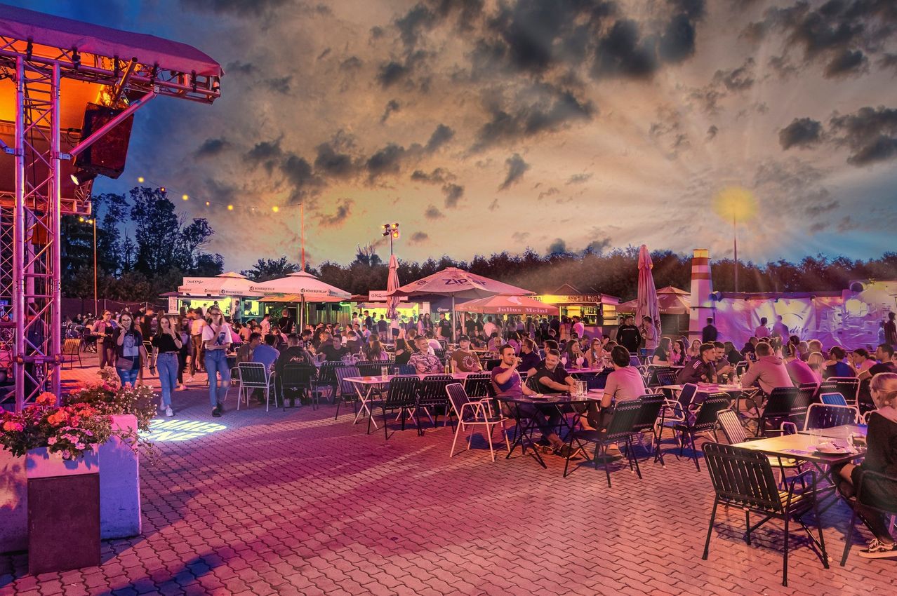 Ein Außenbereich zum Essen bei Dämmerung mit Tischen und Stühlen unter Sonnenschirmen. Menschen sitzen und stehen herum, einige tragen Freizeitkleidung. Der Himmel ist bewölkt mit Rosa- und Lilatönen.
