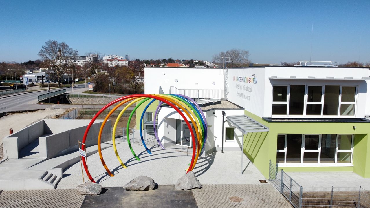 Eine Luftaufnahme eines weißen Gebäudes mit einem bunten Regenbogenbogen davor. Das Gebäude hat ein grünes Dach und ein Glasfenster auf der rechten Seite. Bäume und Gebäude sind in der Ferne sichtbar.