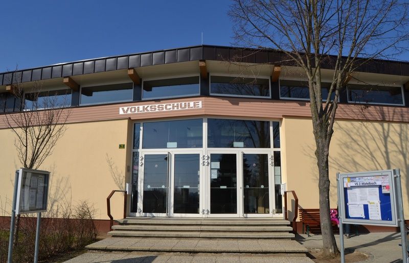 Das Volksschule-Gebäude hat ein modernes architektonisches Design mit einer Glasfassade und einem schwarzen Dach. Es verfügt über mehrere Glastüren mit Handläufen und ein Bulletin-Board mit Informationen steht rechts.