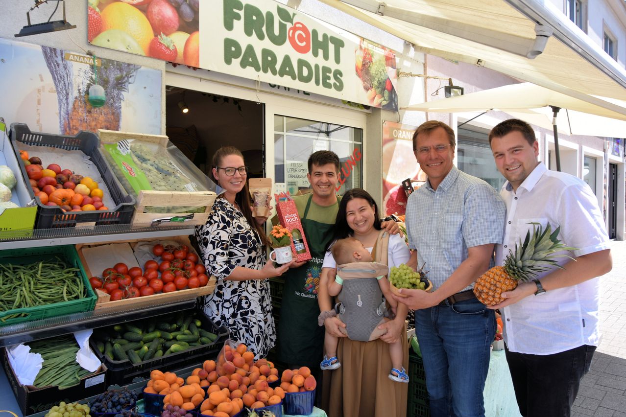 Fünf Personen stehen lächelnd vor einem Obstladen. Sie halten Früchte wie Ananas und Trauben. Der Laden hat ein Schild mit der Aufschrift Fruchtparadiese.