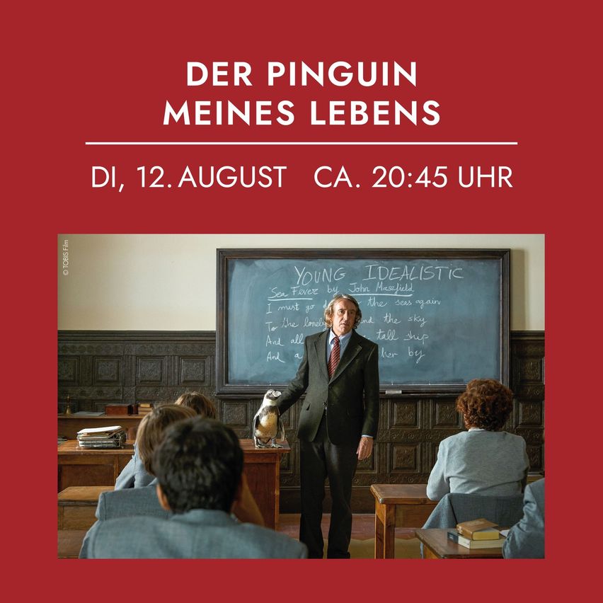 Ein Mann in einem Klassenzimmer hält einen Pinguin. Er steht vor einer Tafel mit Schrift. Die Worte oben lesen 'Der Penguin meines Lebens' und das Datum unten ist der 12. August.