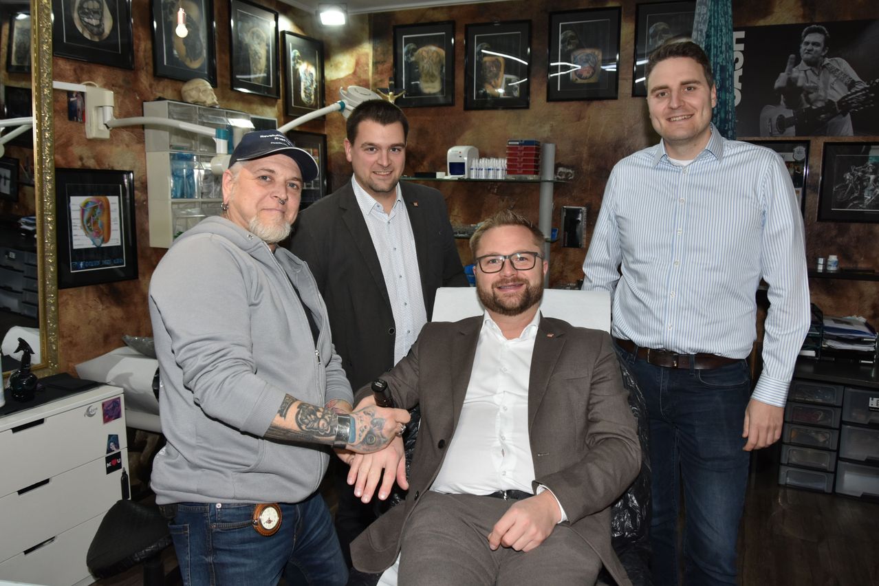 Vier Männer stehen in einem Raum mit verschiedenen Kunstwerken an der Wand. Der Mann in der Mitte sitzt in einem Stuhl, lächelt und lässt sich die Hand halten. Die anderen beiden Männer stehen neben ihm und lächeln.