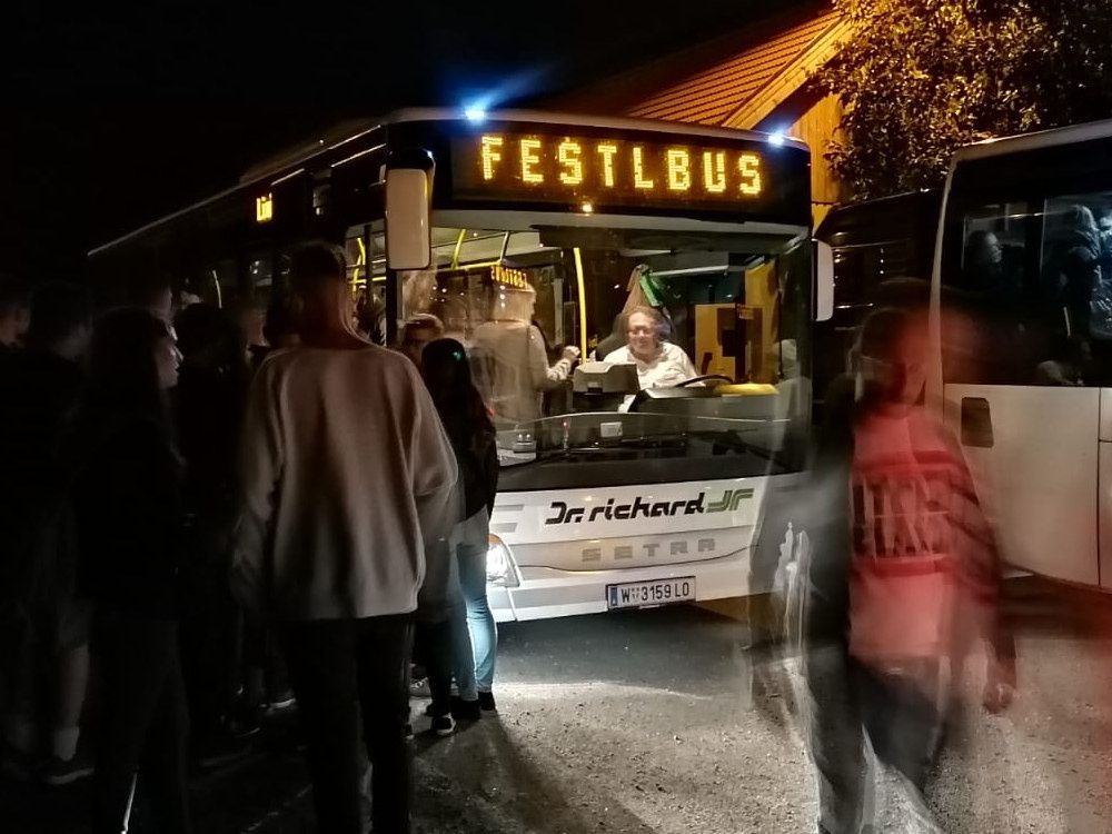 Ein Bus mit dem Schild 'Festlbus' steht nachts geparkt, während Leute einsteigen. Der Busfahrer lächelt und schaut nach vorne.