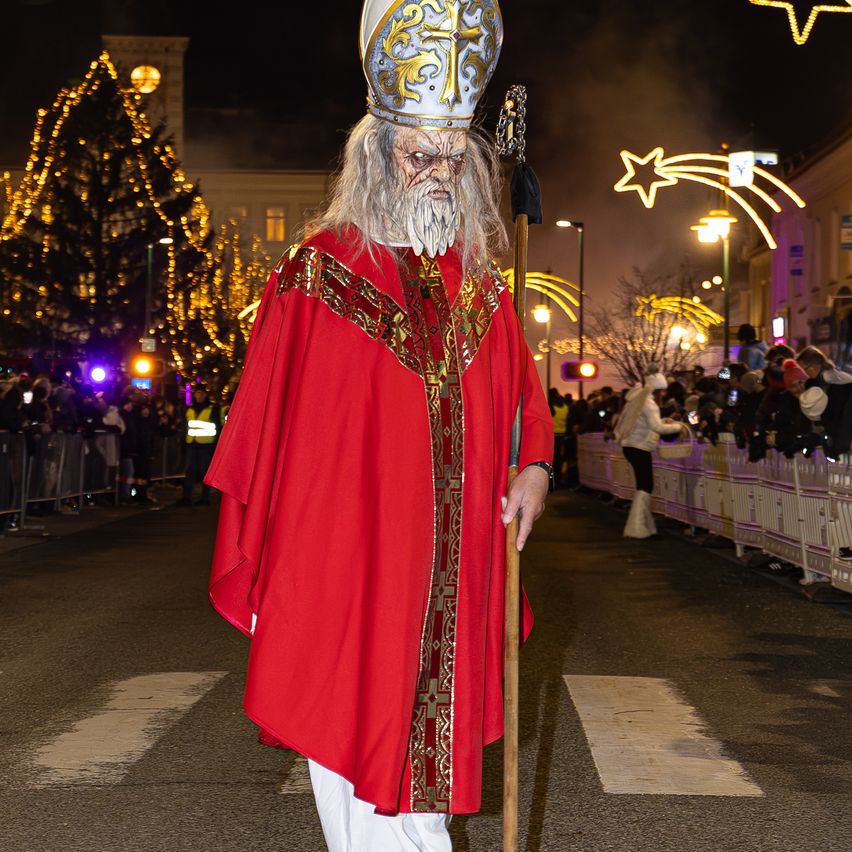 Ein Mann, der als Bischof verkleidet ist, geht bei Nacht auf einer Straße, während eine Menge zuschaut.