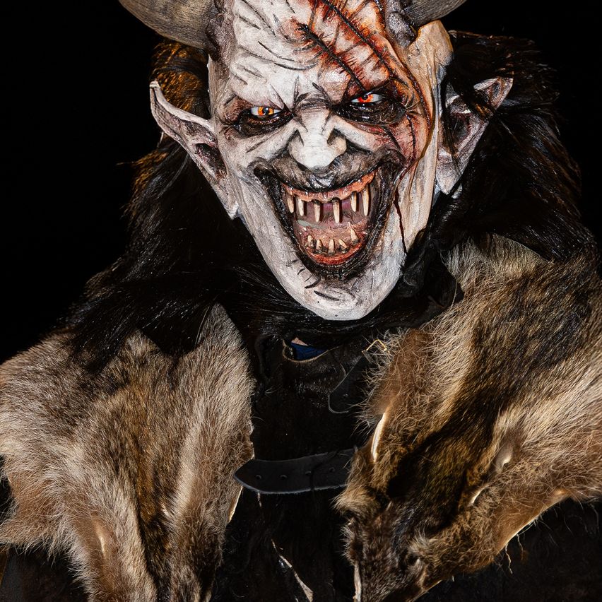 Ein Nahaufnahme eines bedrohlichen Maskes mit Hörnern und einem finsteren Ausdruck. Die Maske ist mit Blut und Schmutz bedeckt und die Person trägt einen Pelzmantel.