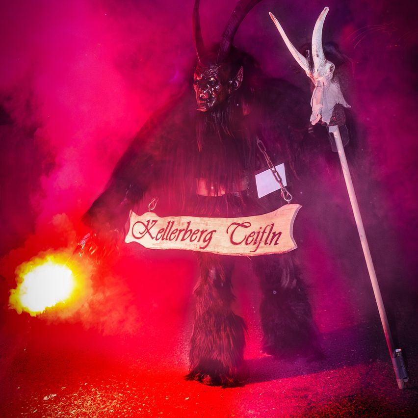 Eine Person in einem Ziegenkostüm mit Hörnern, einem Banner mit der Aufschrift Kellerberg Teufel, hält eine Fackel und einen Stab, mit Rauch im Hintergrund.