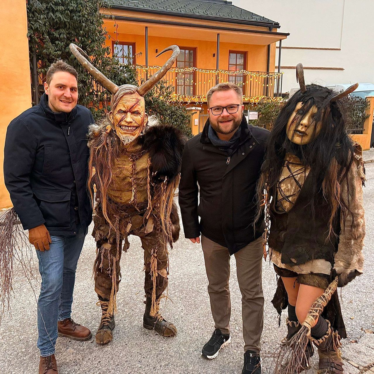 Drei Männer auf einem Foto; einer in einer schwarzen Jacke, ein anderer in einer braunen Jacke und ein dritter in einem gehörnten Monsterkostüm. Dahinter steht ein Gebäude mit Balkon.