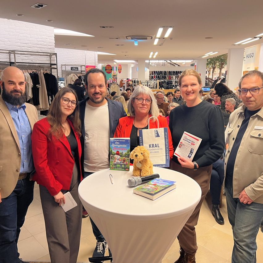 Five people stand around a table in a mall, holding books and a stuffed animal. The table has a microphone and more books. Behind them, people are standing and sitting in the background.
