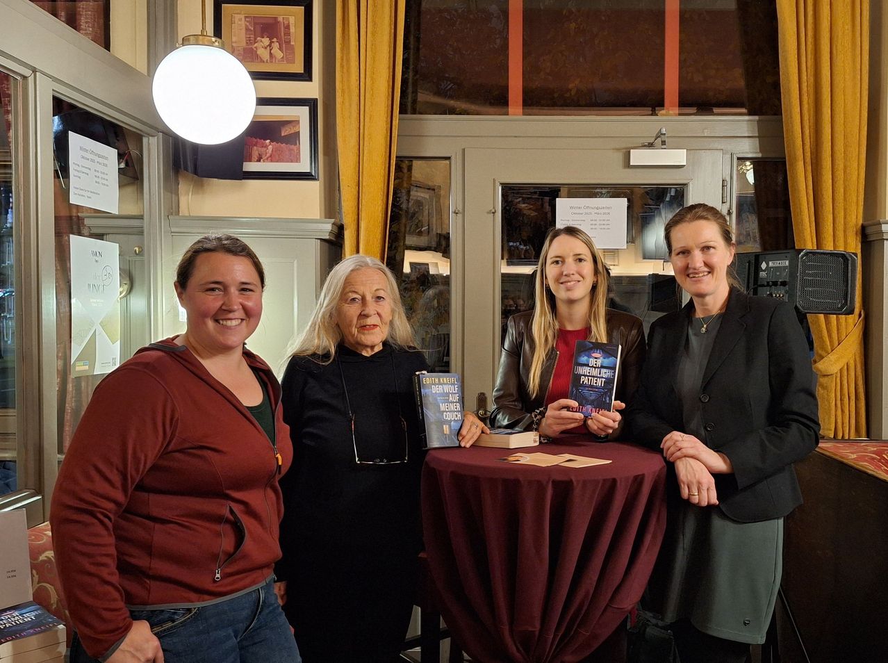 Four women stand around a table, smiling, with books in their hands. The woman on the left wears a maroon hoodie, the second from the left a black dress, the third a leather jacket, and the right a suit. Behind them is a glass door with a sign.