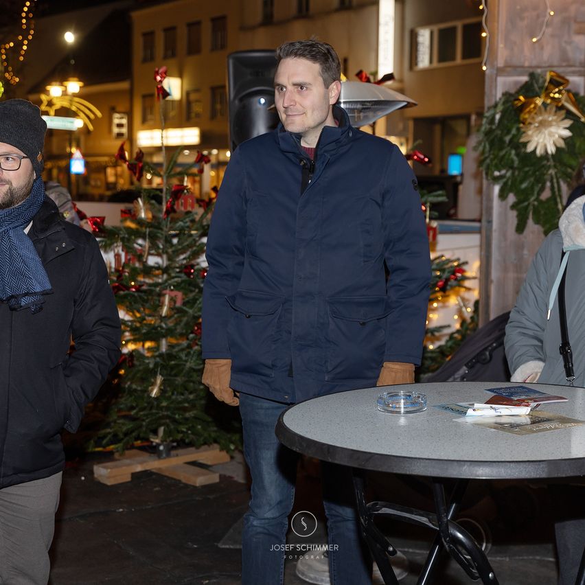 Drei Männer stehen vor einem Tisch mit Broschüren und einem Weihnachtsbaum. Ein Mann lächelt in die Kamera. Im Hintergrund ist eine festlich beleuchtete Straße mit Gebäuden zu sehen.