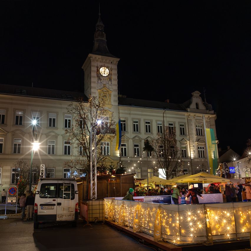 Eine Nachtaufnahme eines Stadtplatzes mit einem Uhrturm. Menschen versammeln sich um eine geschmückte Eisbahn, und ein weißer Lieferwagen ist in der Nähe geparkt. Der Platz ist mit festlichen Lichtern beleuchtet.