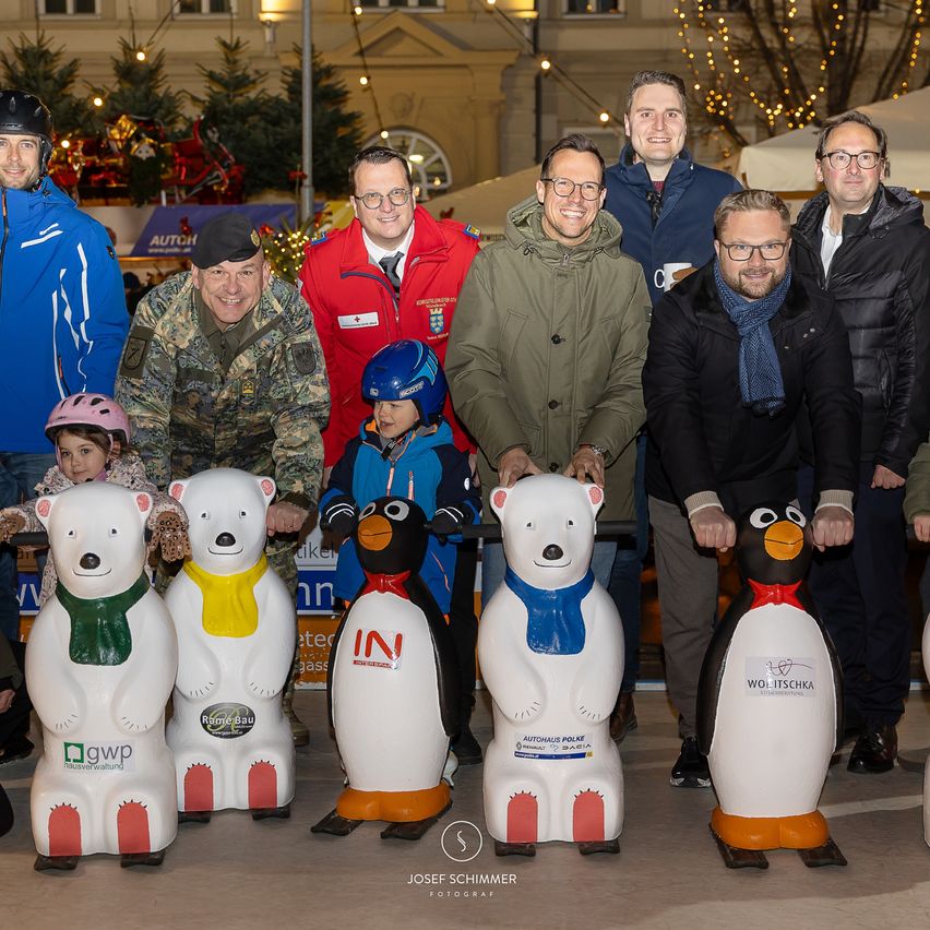 Eine Gruppe von Männern und Kindern steht hinter dekorativen Pinguinen und Eisbären vor einem festlichen Hintergrund und lächelt für ein Foto.