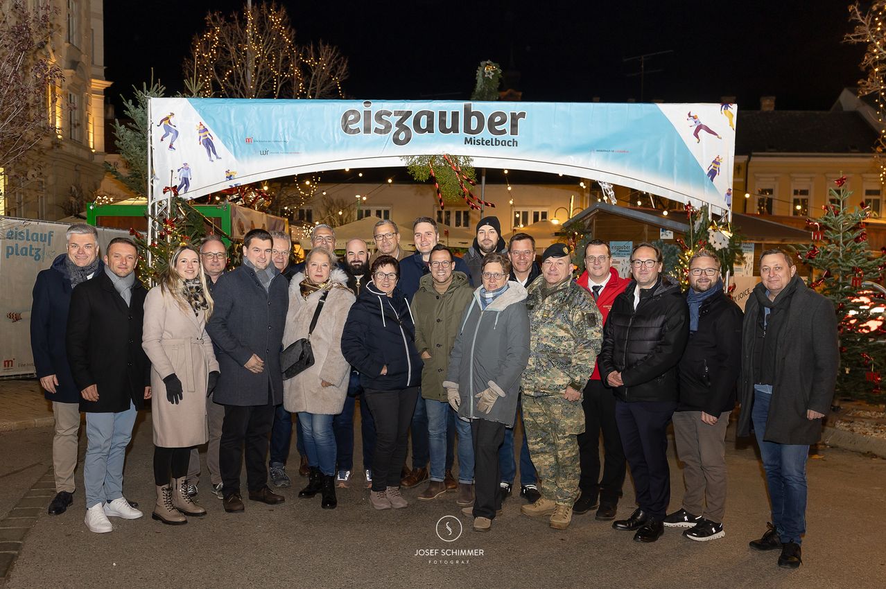 Eine Gruppe von Menschen in Winterkleidung posiert für ein Foto vor einem geschmückten Torbogen, der mit 'Eiszauber Mistelbach' beschriftet ist. Im Hintergrund befinden sich ein Weihnachtsbaum, festliche Lichter und Gebäude.