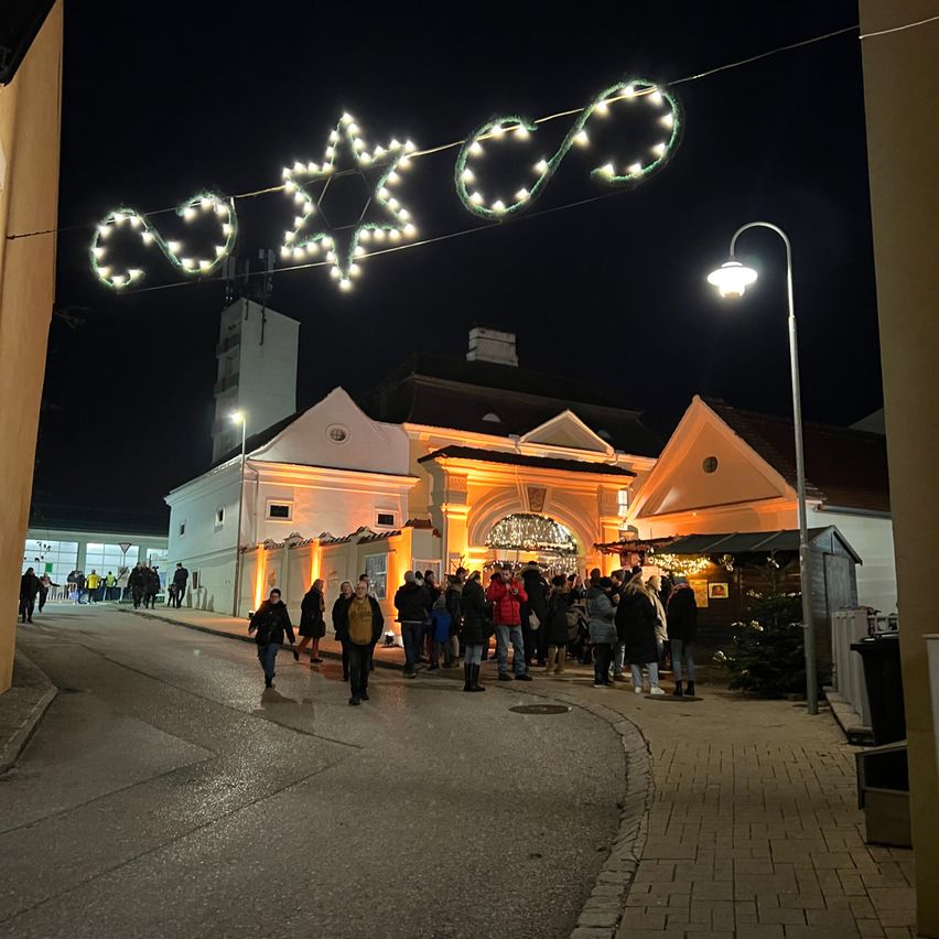 Eine festliche Nachtszene mit Menschen, die an einem Gebäude mit Weihnachtsbeleuchtung vorbeigehen, einschließlich eines Sterns und herzförmiger Lichter.