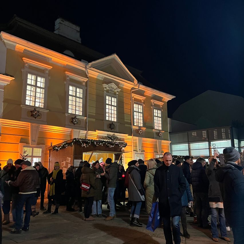 Eine Menschenmenge versammelt sich vor einem warm beleuchteten Gebäude mit Weihnachtsdekoration. Viele Menschen tragen Wintermäntel und Mützen. Einige haben Einkaufstüten dabei.
