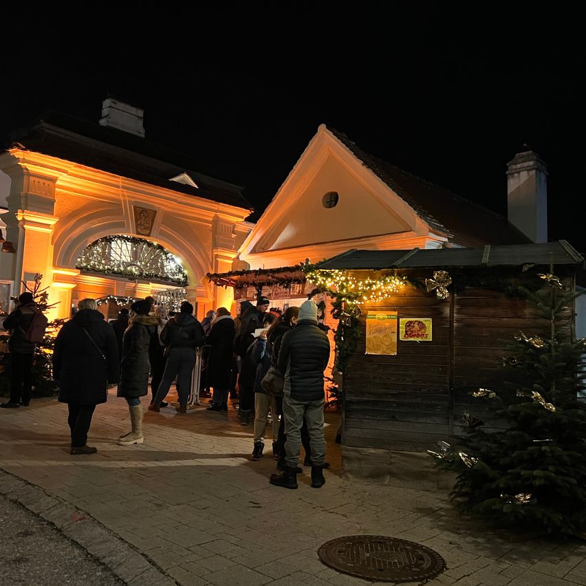 Bei Nacht versammeln sich eine Gruppe von Menschen vor einem Gebäude mit Weihnachtsdekoration, einschließlich eines beleuchteten Baumes und eines kleinen Holzständers mit einem gelben Schild.