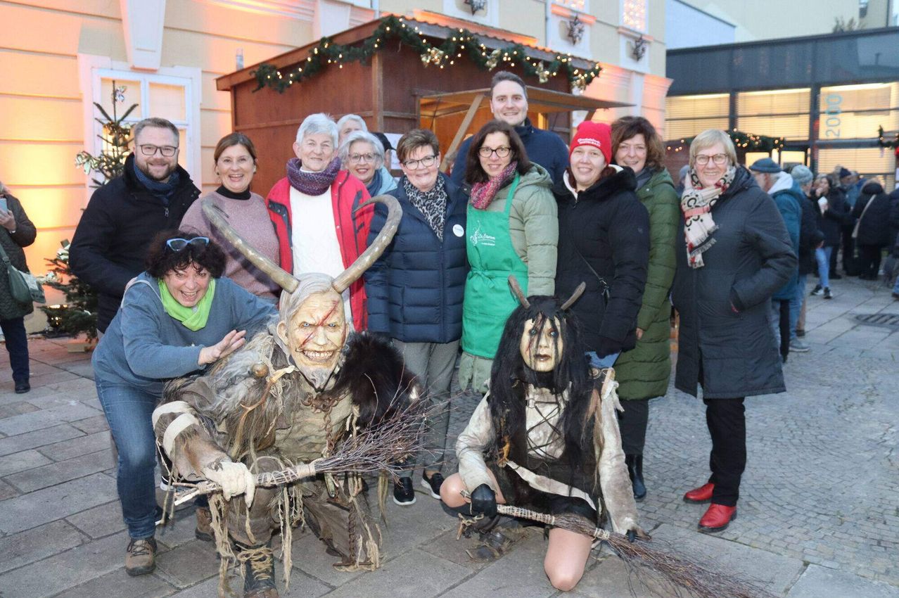 Eine Gruppe von Menschen, einige in Kostümen, posiert für ein Foto im Freien im Winter. Zwei Personen in Kostümen hocken mit Besen, während andere lächelnd um sie herumstehen.
