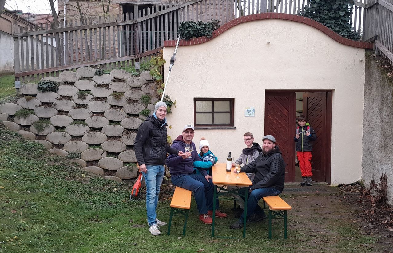 Eine Familie versammelt sich vor einem kleinen Haus mit Tisch und Weingläsern und posiert für ein Foto an einem sonnigen Tag.