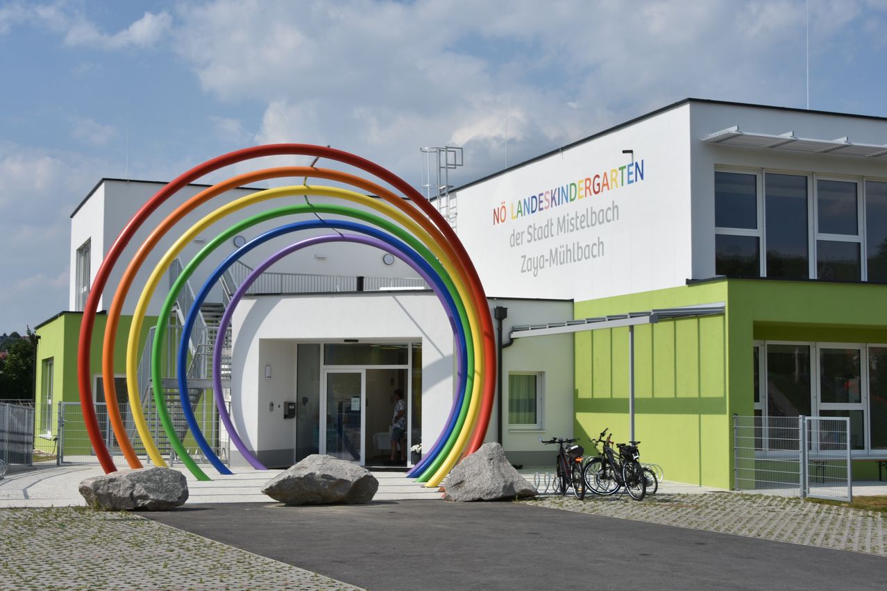 Ein farbenfroher Regenbogenbogen führt zum Eingang des Landeskindergarten der Stadt Mistelbach Zaya-Mühlbach, mit geparkten Fahrrädern an der Seite und einer Person im Inneren.