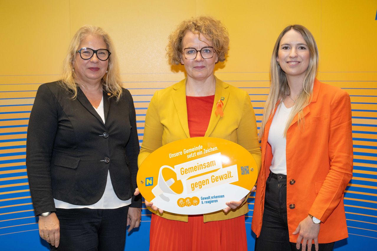 Drei Frauen stehen nebeneinander und halten ein Schild, auf dem 'Gemeinsam gegen Gewalt. Erkennen und handeln' steht. Die Frau in der Mitte hält ein gelbes Schild.