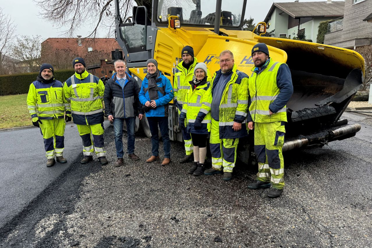Sieben Personen in Warnwesten stehen vor einem gelben Baufahrzeug, wobei eine Frau eine Brille trägt und zwei Männer Bärte haben. Die Umgebung scheint ein Baugelände zu sein.