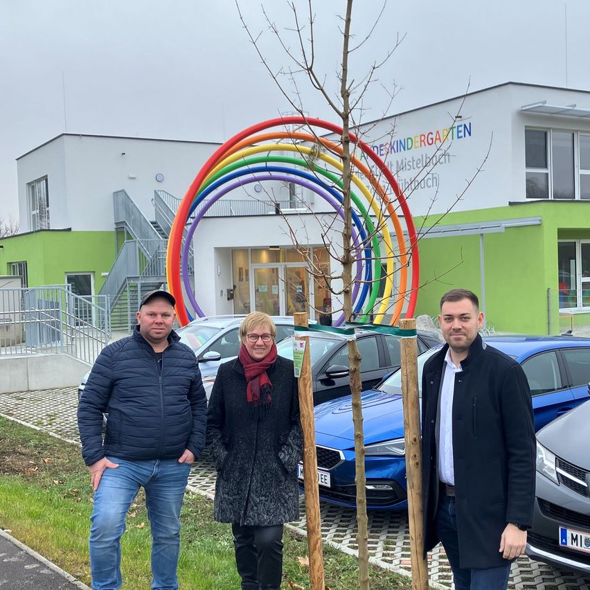 Drei Menschen stehen vor einem Baum und einem Gebäude. Zwei sind Männer und einer ist eine Frau. Sie alle lächeln. Hinter ihnen stehen Autos vor einem Gebäude mit einem Regenbogenbogen und dem Text 'Kindergarten'.