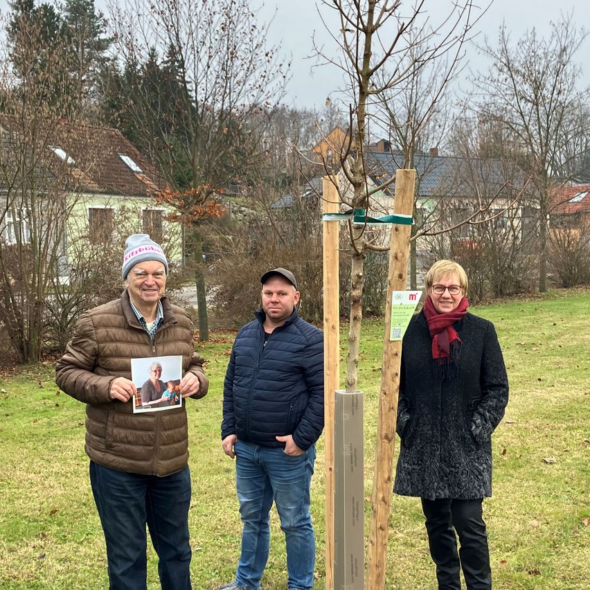 Drei Menschen stehen lächelnd in einem Park. Ein Mann hält ein Foto, ein anderer Mann steht neben einem Baum und eine Frau steht neben einem Holzpfosten. Im Hintergrund sind Häuser und Bäume zu sehen.
