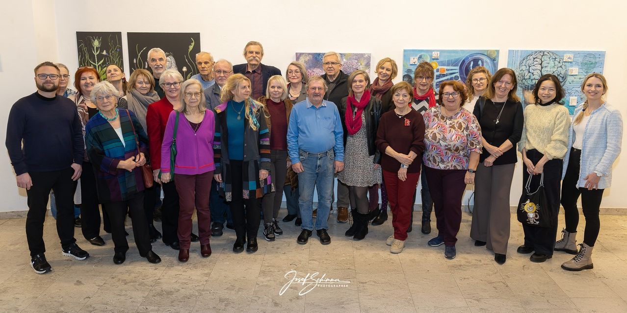 Eine Gruppe Erwachsener in einem Studio, mit Brillen, Halsketten und Schals. Sie posieren für ein Foto vor mehreren Gemälden. Der Boden ist gefliest.