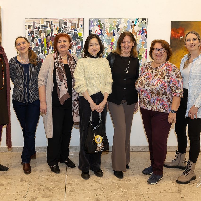 Eine Gruppe von Frauen steht in einer Kunstgalerie und lächelt für ein Foto. Jede hält eine Tasche, und verschiedene Gemälde hängen an den Wänden hinter ihnen.