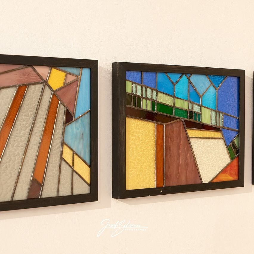 Zwei farbige Glasfenster hängen an einer Wand. Das linke Panel zeigt abstrakte geometrische Formen in Orange, Blau und Gelb. Das rechte Panel zeigt eine Brücke mit einem blauen Bogen, grünen und gelben Paneelen. Beide haben schwarze Rahmen.