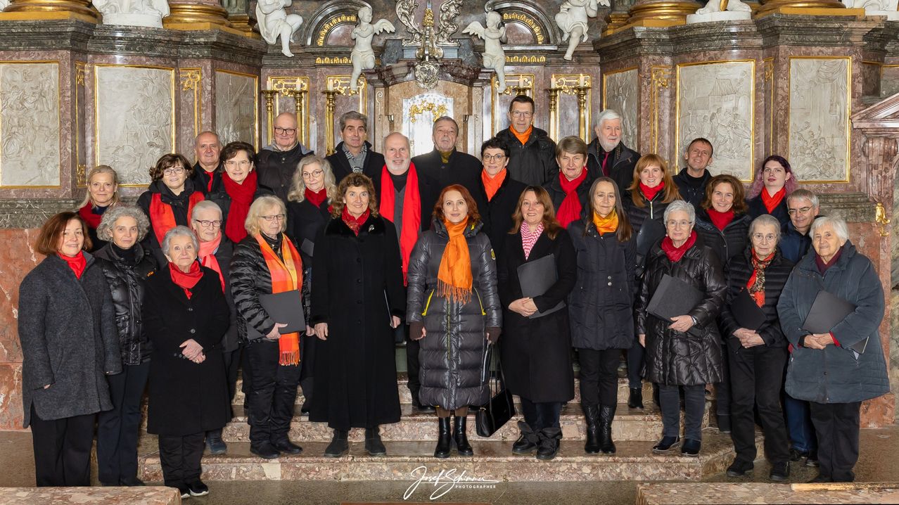 Ein Chor steht vor einem kunstvollen Altar, trägt rote Schals und schwarze Mäntel. Dahinter befinden sich Statuen und ein goldenes Design. Sie halten Ordner in den Händen und posieren für ein Foto.