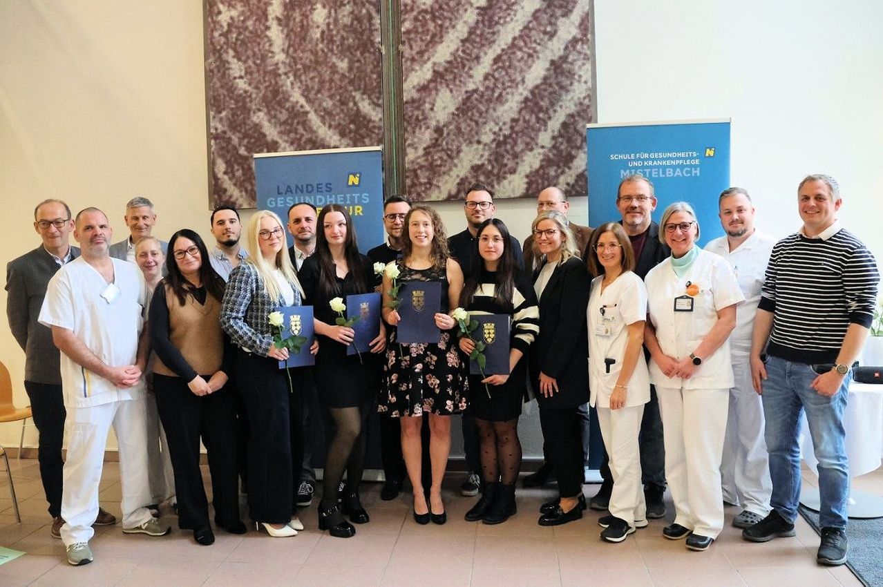 Eine Gruppe von Menschen, darunter Frauen mit Diplomen, steht zusammen in einem Raum. Sie scheinen eine Abschlussfeier oder ähnliches zu feiern. Banner hinter ihnen deuten darauf hin, dass es sich um ein Gesundheitsthema handelt, möglicherweise eine Schule für Gesundheitswesen.