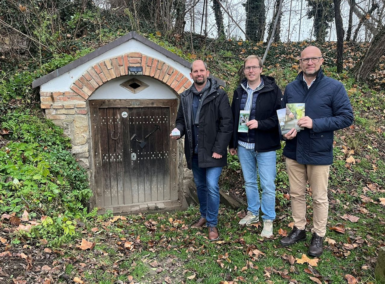 Drei Männer stehen vor einem kleinen, alten Backsteinhaus. Einer hält eine kleine Karte, ein anderer ein Buch und der dritte trägt eine Brille. Sie lächeln in die Kamera.