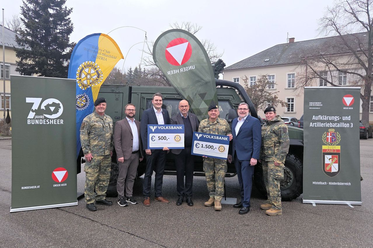 Eine Gruppe von Männern in Militär- und Geschäftskleidung posiert für ein Foto vor einem Fahrzeug und hält einen Spendenscheck für die Volksbank.