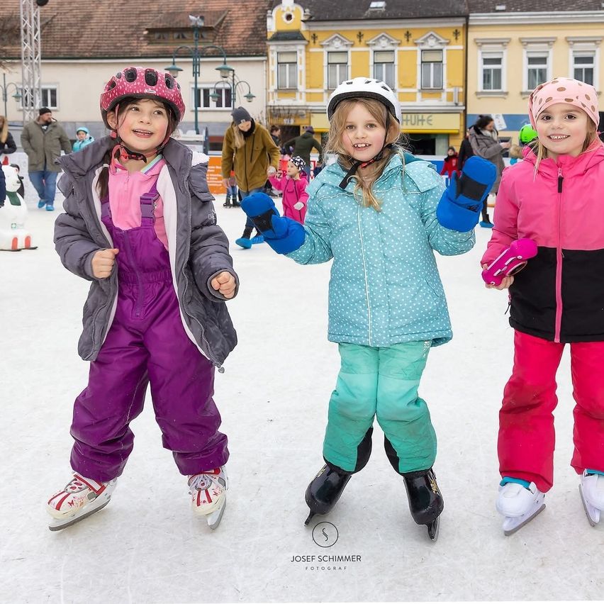 Drei junge Mädchen in Winterkleidung skaten auf einer Eislaufbahn im Freien mit Gebäuden und Menschen im Hintergrund.