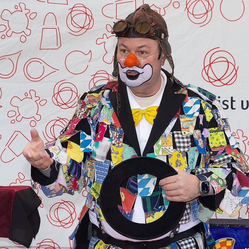 Ein Mann, der als Clown in einem bunten Anzug gekleidet ist, trägt einen Helm, hält einen schwarzen Kreis und gestikuliert mit der anderen Hand. Dahinter befindet sich ein weißer Hintergrund mit roten Kreisen.