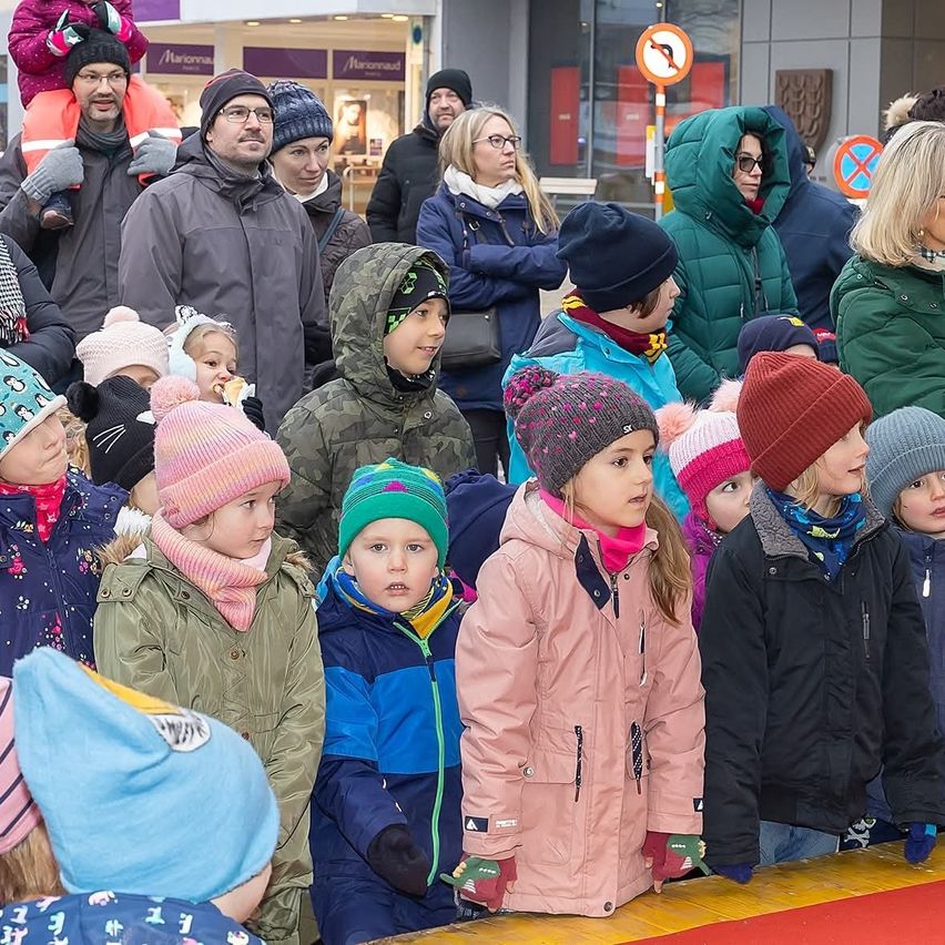 Eine Gruppe von Kindern und Erwachsenen, die warme Winterkleidung tragen, steht in einer Reihe, einige tragen Mützen und Handschuhe, möglicherweise bei einer Veranstaltung im Freien. Im Hintergrund befindet sich ein Gebäude mit Schildern und einer Person, die ein Kind auf den Schultern trägt.