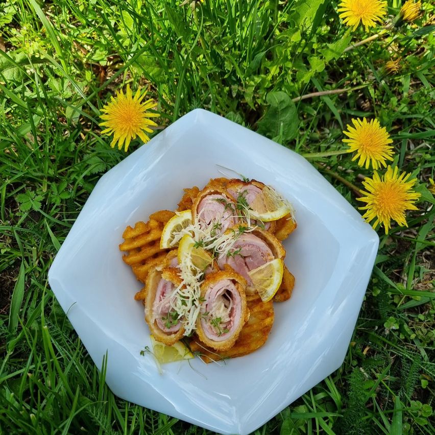 Ein weißer Teller auf Gras hält ein Gericht mit gebratenen Schinkenrollen, garniert mit Zitronenscheiben und Kräutern, umgeben von Löwenzahn.