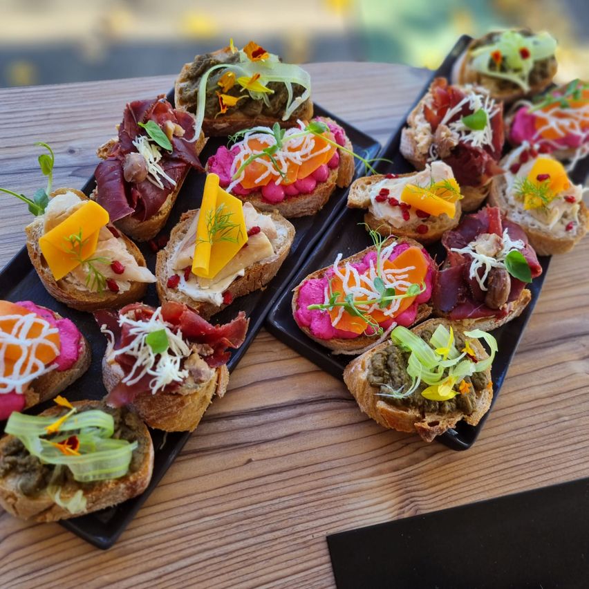 Auf einem Holztisch sind zwei schwarze Tabletts mit Gourmet-Bruschetta zu sehen, jedes belegt mit verschiedenen Zutaten wie Käse, Fleisch und Gemüse, garniert mit Kräutern und essbaren Blumen.
