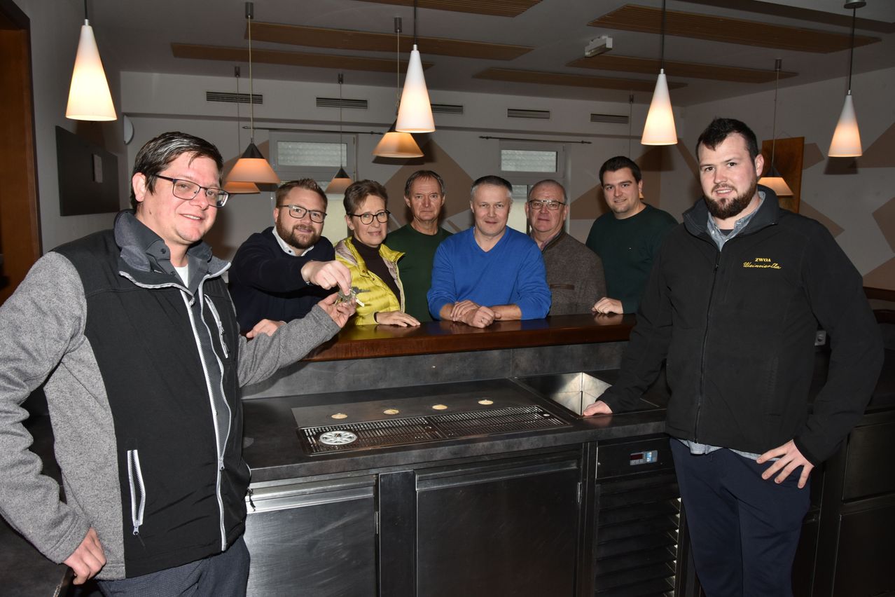 Eine Gruppe von Personen, darunter Männer und Frauen, steht hinter einer Theke in einem Raum. Sie posieren für ein Foto, während einige eine Zigarette halten.