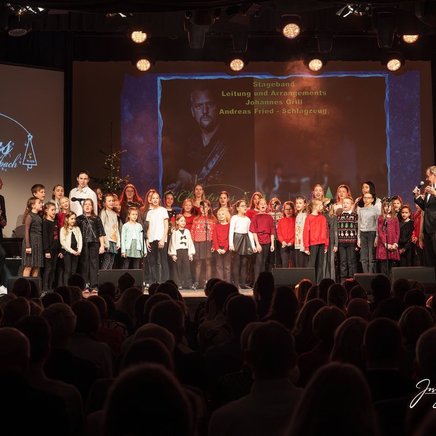 Ein Kinderchor tritt auf der Bühne auf. Ein Mann spricht in ein Mikrofon. Dahinter zeigt eine große Leinwand die Namen der Band und ihrer Arrangements.