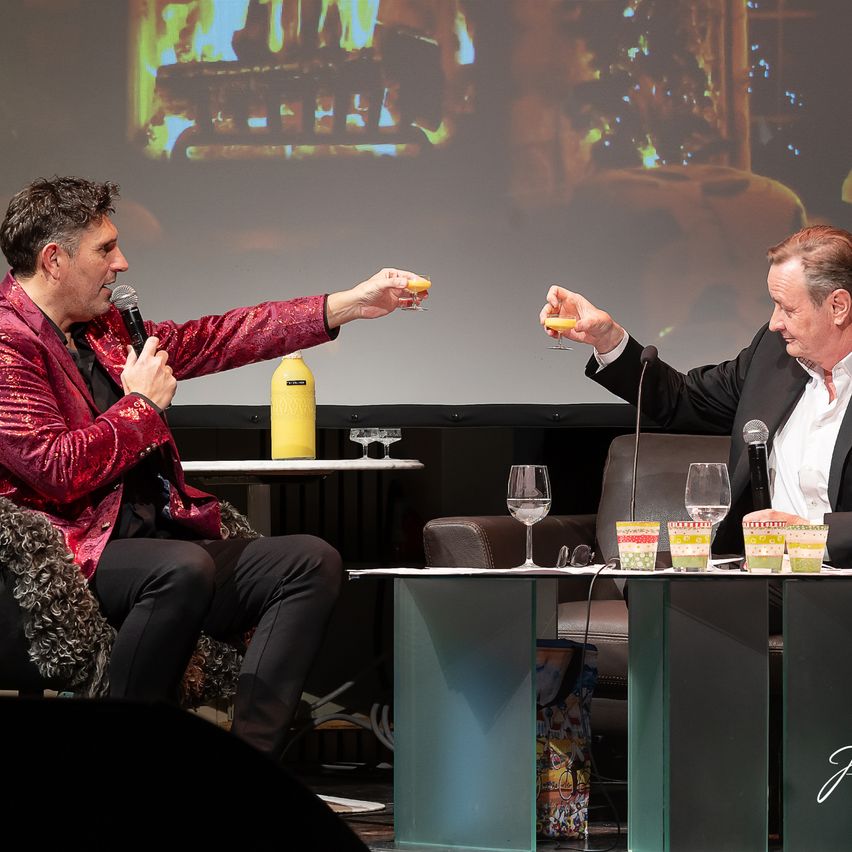 Zwei Männer sind auf der Bühne, einer hält ein Mikrofon, der andere einen Weinglas. Hinter ihnen zeigt eine Projektionsleinwand einen Kamin. Beide Männer sind formell gekleidet.