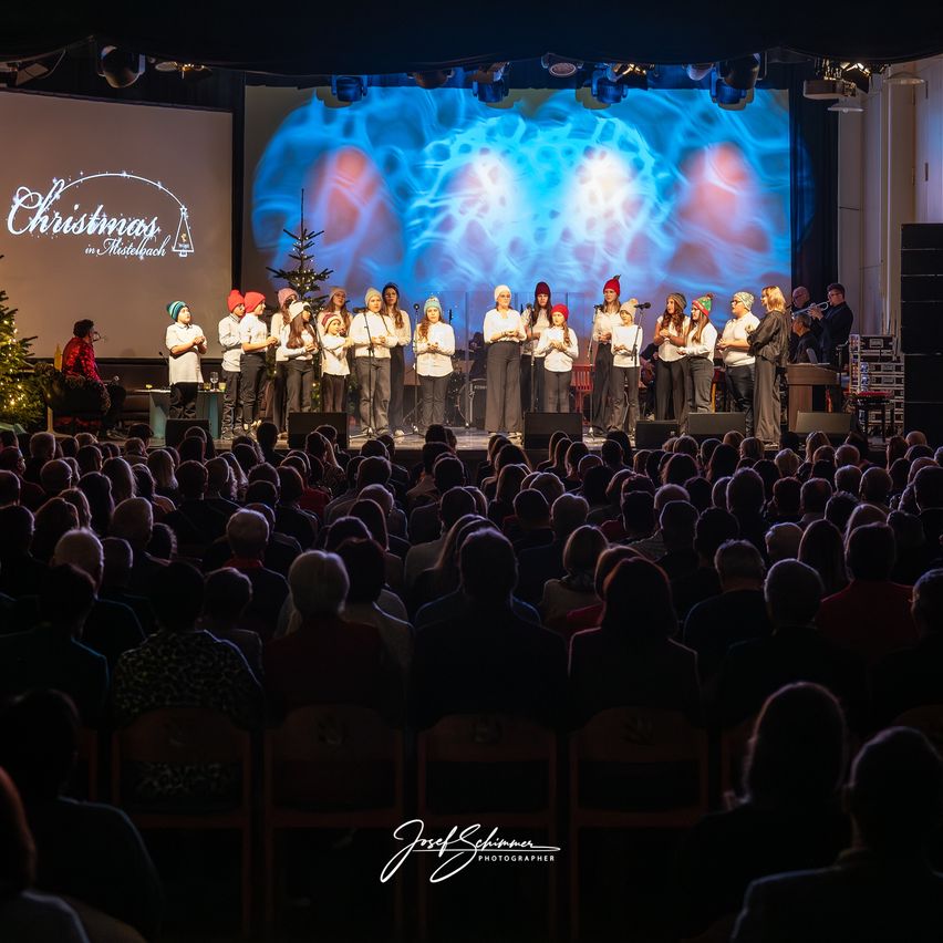 Ein Chor von Kindern in festlicher Kleidung tritt auf einer Bühne auf, auf der eine große Leinwand Weihnachten in Mistelfach zeigt. Ein Publikum schaut von Stühlen zu.