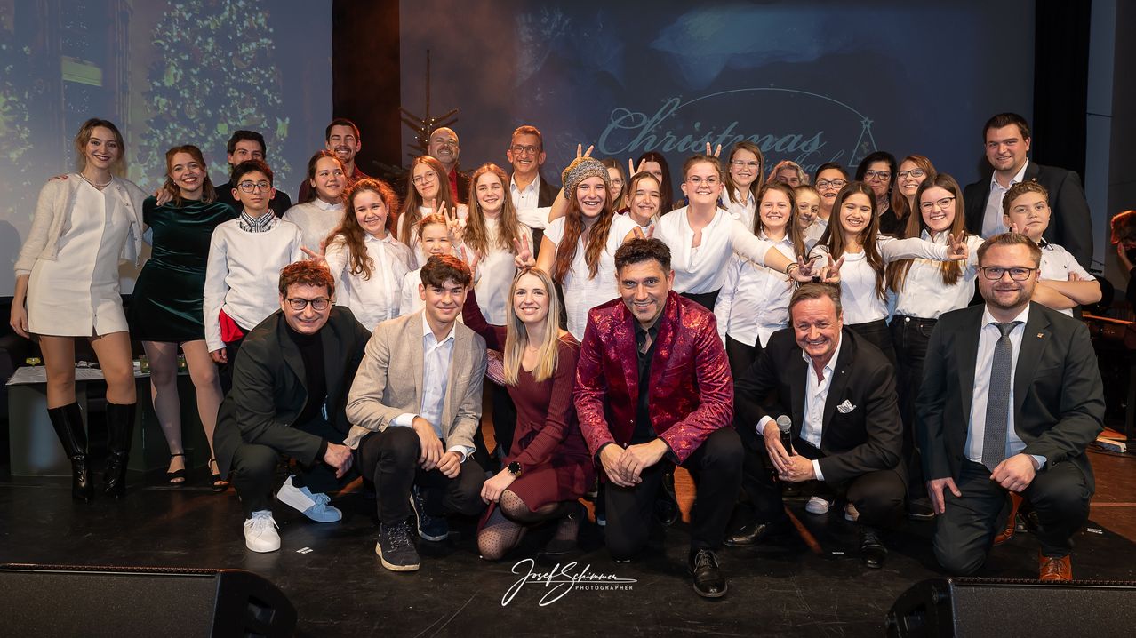 Eine Gruppe von Erwachsenen und Teenagern posiert für ein Foto auf der Bühne. Sie lächeln und zeigen den Friedenszeichen. Ein Mann hält ein Mikrofon.