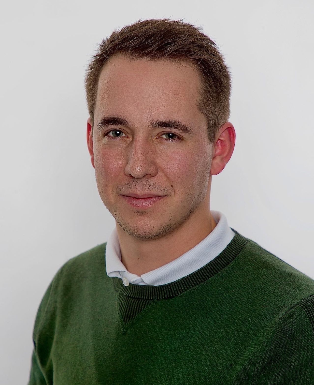 Ein Mann mit kurzen braunen Haaren und einem freundlichen Lächeln trägt einen grünen Pullover und ein weißes Hemd. Er steht vor einem einfachen weißen Hintergrund.