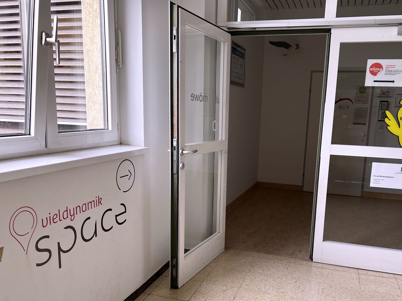 Eine offene Tür mit Glasscheibe führt in einen Raum. Der Raum hat weiße Wände und einen Holzboden. An der linken Wand ist Text geschrieben.