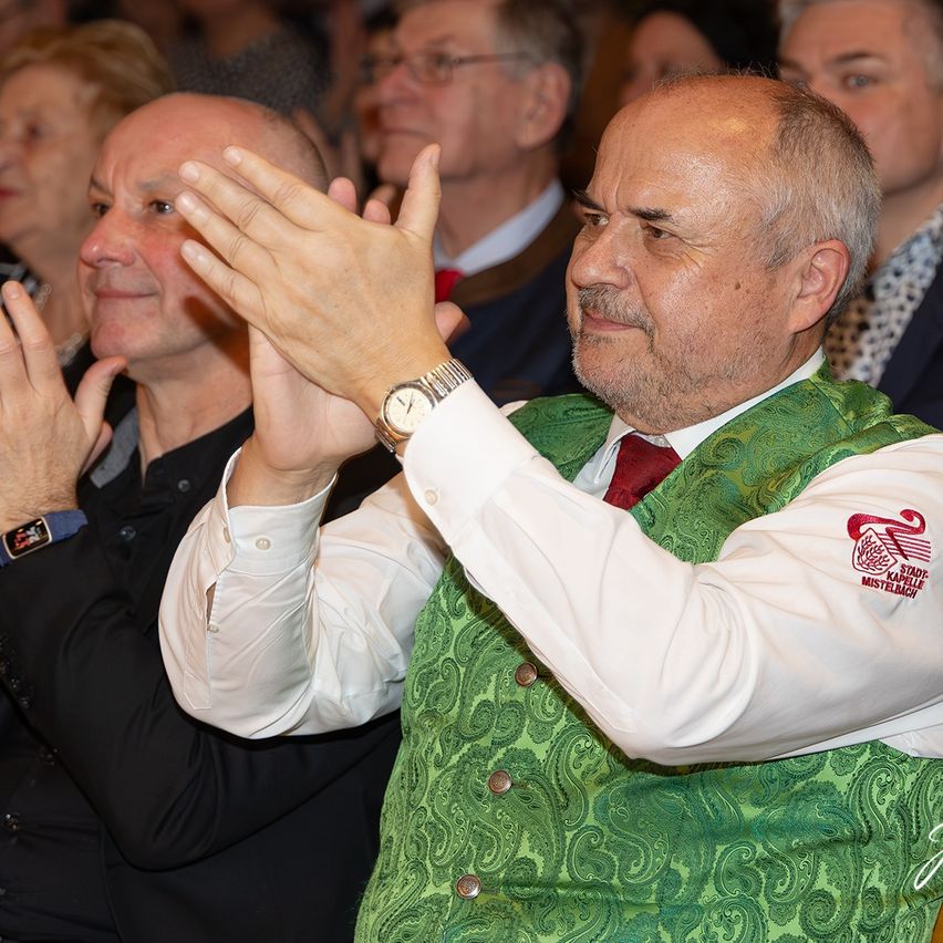 Ein Mann in einem grünen Westchen mit Logo klatscht in einer Menschenmenge. Ein anderer Mann klatscht neben ihm. Sie sind bei einer Veranstaltung.