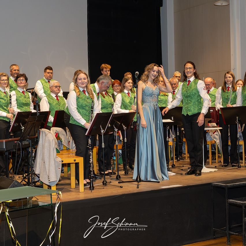 Eine Frau in einem blauen Kleid steht vor einem Chor in grünen Westen und tritt auf der Bühne auf. Sie werden von verschiedenen Musikinstrumenten begleitet, und ein Mikrofon steht in der Nähe.