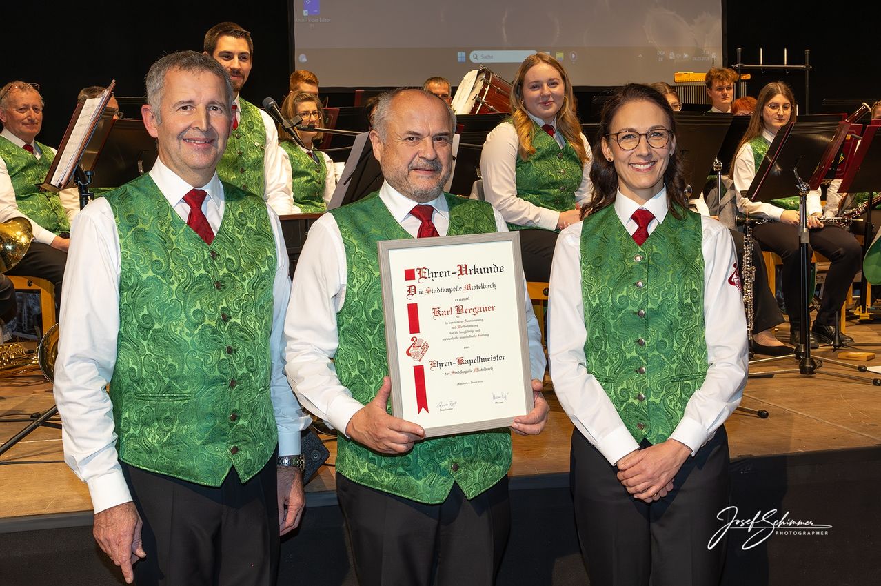 Drei Personen in grünen Westen und roten Krawatten stehen auf einer Bühne, eine hält ein Zertifikat. Hinter ihnen lächelt eine Frau und ein Mann steht mit einem Mikrofon. Im Hintergrund sind eine Trommel und ein Klavier zu sehen.